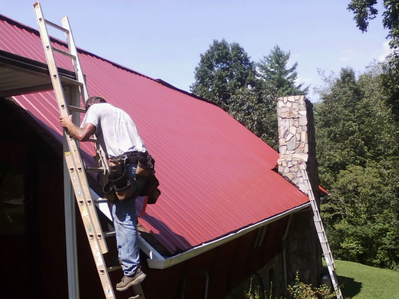 Roofer on a ladder beside a red metal roof and stone chimney — Soffit & Fascia Repair by Roofing Brothers in Sugar Tree